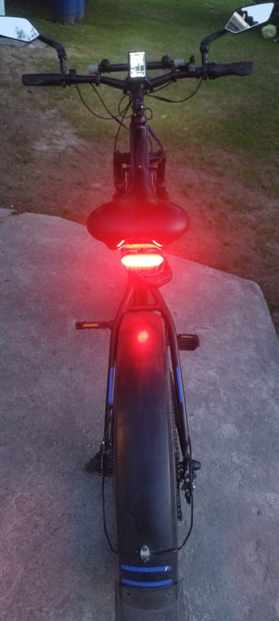 This is a vertical, medium-shot, eye-level, indoor-outdoor image of the rear of a black electric bicycle. The bicycle is parked on a concrete surface, possibly a path or driveway, with a grassy area visible in the background, suggesting a rural or semi-rural setting in Sinkowski Sel'ski Sovet, Belarus.
The bicycle's prominent features include its wide tires, a black saddle with red brake lights illuminated underneath, and handlebars with mirrors. A digital display is visible on the handlebars. The overall lighting suggests it is dusk or nighttime, given the illuminated red lights. The weather appears calm, with no strong indicators of rain or wind.
The image focuses on the parked bicycle, with no people or specific activities evident. Notable details include the bright red glow from the rear lights, the blue reflective strips on the rear fender, and the detail of the bicycle's components like the chain and pedals. The scene is static, with the bicycle appearing stationary. No visible text is present in the image.