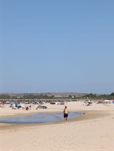 The image depicts a sunny day at a sandy beach in Sines, Portugal. In the foreground, a man wearing black shorts walks towards the viewer across the sand. Behind him, a shallow pool of water with a sandy edge separates him from the main beach area. The main beach is populated with numerous beachgoers, many of whom are sitting under colorful umbrellas. Beach tents and shelters are also visible. In the middle ground, a line of rocks forms a barrier, and beyond that, a hillside is dotted with green vegetation and several wind turbines. The sky is clear and blue.