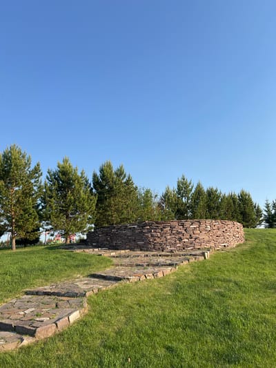 The image depicts an outdoor landscape scene at Емельяновское озеро, Russia, under a clear, bright blue sky that occupies the upper half of the frame. A minute, dark, unidentifiable speck is present in the upper-left quadrant of the sky. The lower half of the image features a vibrant green, well-maintained grassy slope. A path, constructed from irregularly shaped, flat flagstones in varying shades of grey and reddish-brown, winds from the lower-left foreground and ascends towards a stone structure. Grass and small plant growth are visible between some of the flagstones. On the grassy incline, a circular, low wall made of horizontally stacked, flat, reddish-brown and grey stones is prominently featured. This structure appears to be a dry-stone construction. Behind the stone structure and extending across the mid-ground, a line of tall, coniferous trees, consistent with pine trees, stands with dense green foliage. Through the gaps in these trees on the left side of the image, glimpses of brightly colored elements, suggesting playground equipment, are faintly discernible in the distant background. The scene is illuminated by direct daylight, casting an even brightness across the environment. There are no individuals or identifiable animals present in the frame.