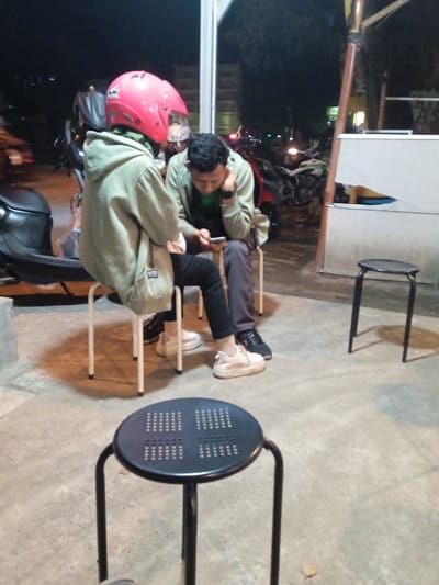 This image captures two individuals seated on simple metal stools at what appears to be an outdoor or semi-outdoor casual gathering spot at night in Sekopek Wetan, Indonesia. The ground is concrete, and artificial lighting illuminates the scene, casting shadows.
The person on the left is seen from behind, wearing a prominent pink motorcycle helmet with "INK" visible on the side. They are dressed in a light green or khaki hoodie over a green inner layer, black trousers, and white sneakers. To their left, a black scooter is partially visible.
The second individual sits facing partly towards the camera, looking down at a smartphone held in their hands. They are also wearing a similar light green or khaki hoodie over a green top, dark trousers, and black shoes. Their expression is focused on the phone.
In the immediate foreground, a black metal stool with a perforated circular top is positioned, acting as a table or an unoccupied seat. Another black stool stands unoccupied in the mid-ground on the right. In the background, several motorcycles are parked, and faint lights from other establishments or vehicles are visible in the dark surroundings. The overall setting suggests a relaxed, informal social interaction during the evening.