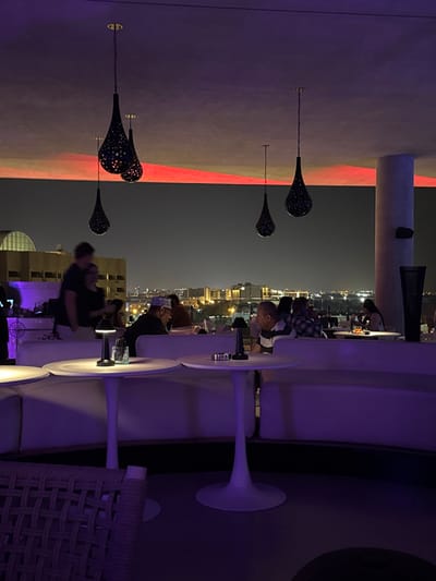 An elevated outdoor seating area at night with city views. White tulip tables and cushioned seating are visible, some occupied by people. Overhead, decorative teardrop-shaped light fixtures adorn a wall accented with red light. The urban landscape twinkles in the background, suggesting a rooftop or high-rise setting. Soft, purple lighting enhances the modern and relaxed ambiance.