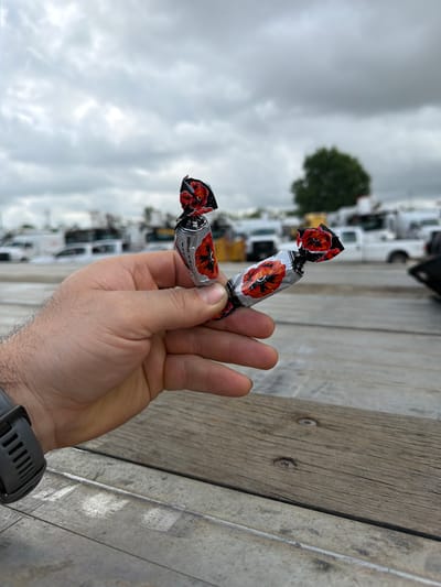 A close-up, vertically oriented outdoor shot features a person's hand holding two wrapped candies against a blurred background. The hand, with visible hair on the wrist and a dark gray smartwatch, extends from the lower right of the frame, holding the candies in the center. The candies are individually wrapped in silver foil with a distinctive red poppy flower design, black accents, and green stems. Visible text on the wrapper includes "КРАСНЫЙ ОКТЯБРЬ" (Krasny Oktyabr - Red October) and "МАК" (Mak - Poppy), along with smaller English text "POPPY SEED PRALINE."

The immediate foreground features a worn, light brown wooden plank secured by two visible screws, next to what appears to be a concrete or metal beam. The background is heavily blurred but reveals an outdoor setting. Numerous white commercial vehicles, possibly vans or trucks, are parked in rows, suggesting a parking lot or industrial area. A single, dark green deciduous tree stands out against the muted tones of the sky and vehicles. The sky is overcast with gray clouds, indicating a cloudy daytime. No specific human emotions are discernible. The location is an unspecified outdoor area within Midlothian, United States.