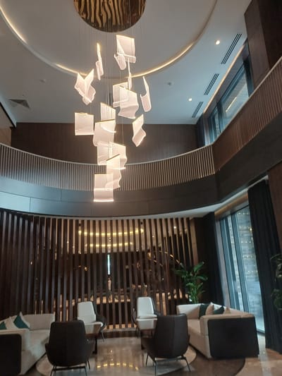 The image depicts the interior of a modern, upscale lobby or waiting area, likely in a hotel or commercial building. The scene is dominated by a large, multi-tiered contemporary chandelier featuring numerous translucent, cube-like elements suspended by thin wires from a circular, metallic ceiling fixture.
The architecture of the space is characterized by curved walls and multiple levels, indicated by a balcony with a white railing visible in the mid-ground. The walls are finished with dark wood paneling and contrasting dark brown wall coverings. The flooring appears to be polished marble or a similar reflective material.
Furnishings include several comfortable seating arrangements. On the left, a light-colored sectional sofa with turquoise accent pillows is partially visible. In the foreground, a dark charcoal armchair with a metallic base is prominently featured. To the right of this, a lighter-colored armchair with a dark base is positioned near a small table. Further back, another light-colored sofa with dark green pillows is situated, accompanied by large potted plants that add a touch of greenery to the sophisticated ambiance.
Large windows in the background reveal a glimpse of the exterior, possibly showing other buildings or city lights, suggesting an urban setting. The overall impression is one of contemporary elegance and spaciousness.
The location in the city of Myk, Uzbekistan is not discernible from the visual evidence provided in the image.