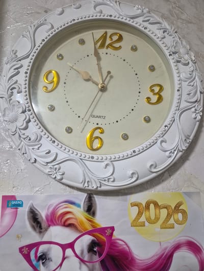 This media file displays a close-up view of a decorative white wall clock with a cream-colored face. The clock features gold-colored Arabic numerals for 3, 6, 9, and 12, with smaller gold-colored accent numbers at other positions. Several small, clear rhinestones are embedded in the clock face. The clock hands are gold-colored and appear to be pointing close to 9:22. The clock has an ornate, embossed frame with floral and leaf motifs. To the right of the clock, a poster is partially visible. The poster shows a cartoon-like image of a unicorn or horse with a rainbow-colored mane and wearing pink cat-eye glasses. A circular emblem on the poster contains the text "2026" in large gold glitter letters. Below this, the poster has text in Cyrillic script which reads "QAZAQ БРЕНД Типография". The setting appears to be indoors, likely a room in Қанабек, Kazakhstan, given the provided location context. There are no visible indications of time of day, weather, or strong emotions conveyed by the image. The overall impression is of a decorative item and a piece of promotional material.