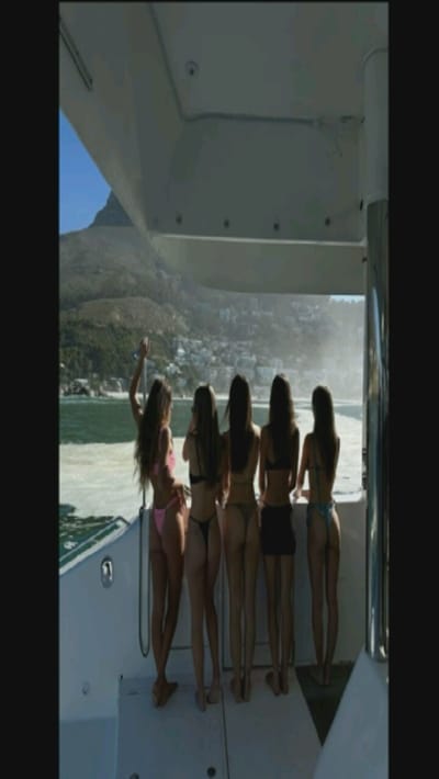 Five women stand on the stern of a boat, facing the sea and distant hills. They wear swimwear and varying shades of dark hair. The boat is white with metal supports. A portion of a mountainous landscape is visible across the water, slightly obscured by the bright haze. The scene is sunlit.