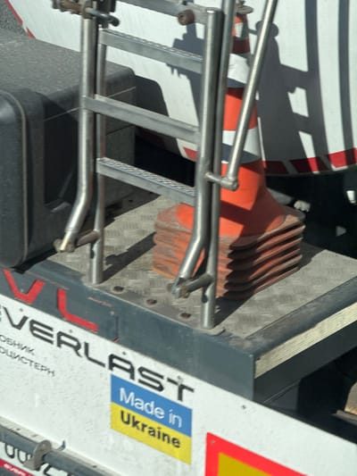 The image displays a close-up, elevated view of the rear or side platform of a utility vehicle, situated on Peremohy Street, Ukraine. The platform itself is constructed from metallic diamond plate. A polished metal ladder with multiple rungs is securely mounted on this platform. To the right of the ladder, a stack of at least six orange traffic cones, each featuring white reflective bands, is visible, with the uppermost cone fully exposed. A dark, rectangular object, possibly a storage box or toolbox, is partially visible on the platform's left side. Directly below the platform, on the vehicle's white body, the brand name "EVERLAST" is printed in black lettering, with the "V" in red, accompanied by additional, partially visible Cyrillic text. Below the "EVERLAST" branding, a blue and yellow sticker, resembling the Ukrainian flag, prominently displays the text "Made in Ukraine." A partial red and yellow reflective marker is visible at the bottom right of the frame. Strong, defined shadows are cast across the white vehicle surface and the platform, indicating bright, direct sunlight. No individuals are present in the scene.