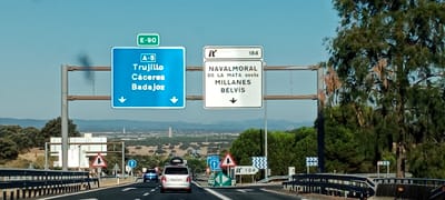 A sunny day scene captures a highway with directional signs in Millanes, Spain. In the foreground, a grey vehicle with a rooftop cargo carrier is driving on a highway. To the left, a blue car is also traveling on the highway. The road is flanked by safety barriers and trees. Overhead, two large blue and white signs provide directions. The blue sign on the left indicates "E-90", "A-5", "Trujillo", "Caceres", and "Badajoz" with downward arrows. The white sign on the right shows "Navalmoral De La Mata Oeste", "Millanes", and "Belvis" with a downward arrow and the number "184". In the background, a vast landscape with rolling hills and scattered trees stretches to the horizon under a clear blue sky. Several distant structures, possibly towers or buildings, are visible in the hazy distance.