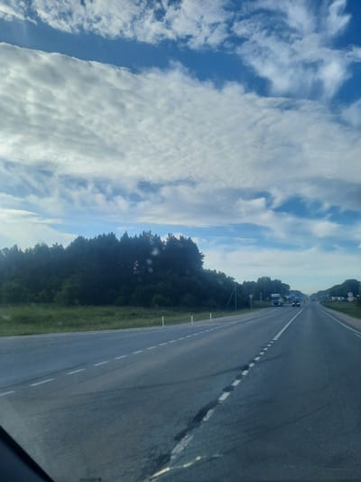The image shows a highway in the city of Pyatiletka, Russia. The sky is filled with dramatic clouds, and the road is lined with trees. There are a few cars and trucks on the road. The image is taken from the perspective of someone in a car, looking forward.
