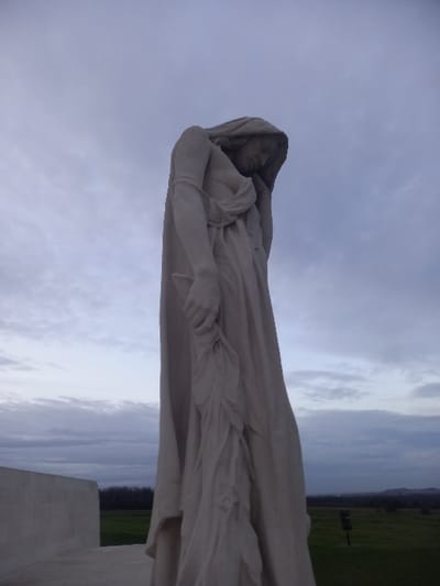 A white marble statue of a draped female figure is set against a sky of gray clouds. The figure is in repose, possibly sleeping or deceased. The statue is positioned outdoors, with a glimpse of green fields and trees in the background. The figure's face is serene, with a hooded drape partially covering her head. Her hands are gently clasped together, holding what appears to be a wreath.