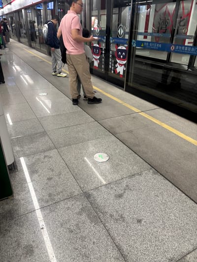 A subway station scene. People stand on a gray tile platform near a train. Yellow warning lines mark the platform edge. Safety instructions are visible on the train doors, written in Chinese and English, with pictograms showing safety precautions. A round floor sign indicates the direction of movement. The lighting is bright, and the scene has a clean, modern appearance.