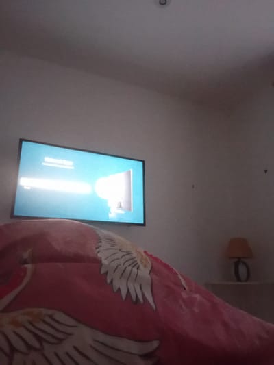 A television screen displays a bright light on a light blue background with text partially visible. The television is mounted on a wall in a room. In the foreground, a pink blanket with a white bird pattern covers a surface, possibly a bed. To the right of the television, a small lamp with an orange shade sits on a shelf. The overall lighting in the room appears dim. The scene is indoors and appears to be a bedroom setting. There is no visible indication of the city of Portree, United Kingdom in the image.