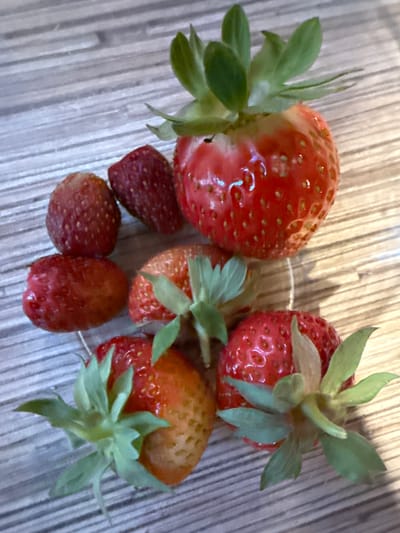 A close-up, overhead shot shows seven strawberries on a wooden surface. The strawberries are red and vary in size, with some having green leafy tops. The wooden surface appears to be a table or countertop with visible grain lines. The lighting suggests an indoor setting, possibly a kitchen. The image is focused on the fruit, with no indication of human presence or activity. The location is specified as Петрищево, Russia.