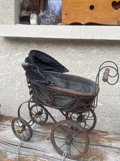 The image shows a vintage doll-sized baby carriage with a black fabric hood, a wicker-like basket, and wooden wheels. It is placed on a rustic wooden surface in front of a textured wall. In the background, on a white ledge, are several decorative items: a brown, bird-shaped object, a small owl figurine with large eyes, and a snail figurine decorated with colorful stones. The overall setting appears to be outdoors, possibly on a windowsill or a porch, in a location that evokes a sense of quaintness or rustic charm. The city of Les Rousses, France, is a mountain resort town known for its natural beauty and traditional architecture, which aligns with the aesthetic of this image.