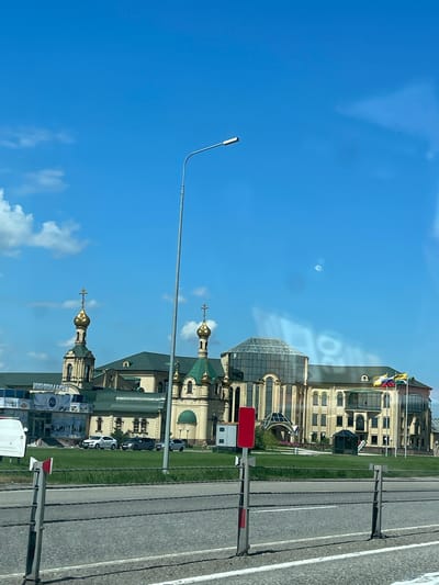 An eye-level, exterior medium shot captures a modern complex with traditional architectural elements under a clear blue sky with scattered clouds. The building features multiple domes with golden crosses, characteristic of Russian Orthodox churches, and a large, modern glass-domed section. The facade is a pale yellow with arched windows. In front of the building, a manicured green lawn is visible, with a few cars parked on a paved area. Flags are flown on poles to the right of the building. A street light stands tall in the foreground, to the left of center. A divided highway runs across the bottom of the frame, with a metal barrier separating it from the lawn. The scene is located in Ul'yanovka, Russia.