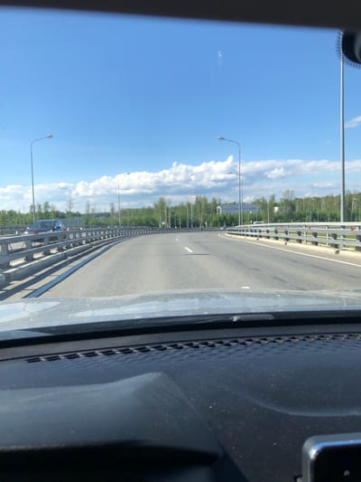 The image captures a wide-angle view from inside a vehicle traveling on a multi-lane highway or bridge on a clear, sunny day in Levashovo, Russia. The perspective is from the passenger side, with part of the dark dashboard or car interior visible on the right and a sliver on the top left.

The asphalt road stretches into the distance, marked with white dashed lines indicating lanes. Sturdy metal guardrails line both sides of the highway, separating it from the surrounding environment. Beyond the guardrails, dense green trees and foliage are visible, suggesting a wooded or forested area.

Numerous tall, slender lamp posts with curved tops line both sides of the road, evenly spaced and receding into the horizon. In the distance, on the left side of the road, another vehicle, possibly a dark-colored car, can be faintly seen behind the guardrail.

The sky is a vibrant blue, mostly clear but with scattered fluffy white clouds, particularly along the right side of the frame. The bright sunlight casts distinct shadows, especially from the guardrails onto the road. The overall impression is one of travel and open highway on a pleasant day. No discernible text is visible anywhere in the image.
