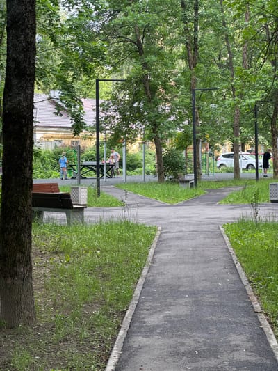 The image depicts an outdoor scene with a paved pathway in the foreground that splits into two diverging paths. The pathway is flanked by grassy areas and trees. In the mid-ground, several people are visible in what appears to be a park or playground area. One man and two children are near a table, possibly engaged in a game. Another individual, dressed in dark clothing, stands near a tree on the right side of the image. A white car is parked in the background, with buildings and more trees surrounding the area. The scene suggests a residential or community space in Kuzmolovskiy, Russia, with elements of recreation and everyday life.