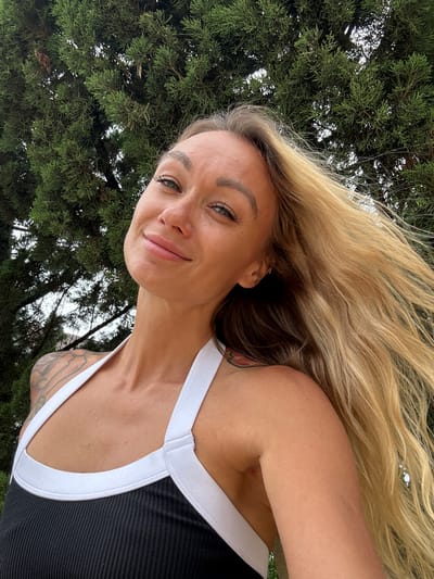 This is a selfie of a woman with long, blonde, wavy hair. She is wearing a black tank top with white trim. Her head is tilted to the side, and she is smiling at the camera. Her left arm is bent and resting on her head. There are tattoos visible on her left shoulder and upper arm. The background is out of focus, but appears to be a dense green foliage, possibly trees, with glimpses of a cloudy sky. The image has a natural, outdoor feel. No text is visible.