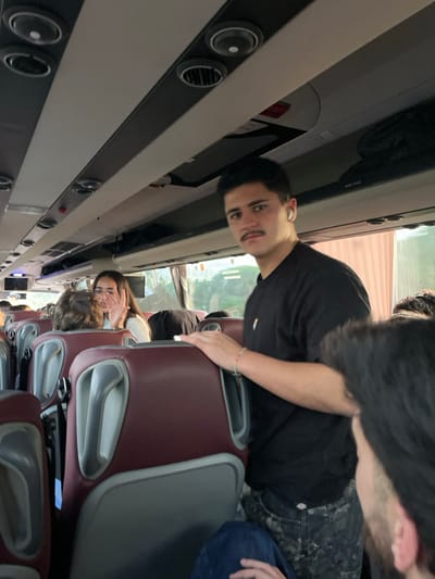 The image depicts the interior of a passenger coach or bus. The location within the city of Leiria, Portugal, cannot be definitively determined from the visual evidence.
In the mid-ground, a young male stands facing the camera, positioned slightly right of center. He has dark hair, a mustache, and is wearing a black t-shirt with a small colorful logo on the left chest, and dark patterned trousers. White earbuds are visible in both ears. His right hand rests on the upper part of a burgundy and grey bus seat. He wears a light-colored beaded bracelet on his left wrist. He looks directly at the camera with a neutral expression.
Behind this male, to the left, a young female with dark hair and glasses is seated, looking towards the camera. Her right hand is raised, palm facing the camera, in what appears to be a gesture to stop or block the image capture. She is wearing a light-colored top.
In the immediate foreground, the back of a person's head and shoulder with dark hair is visible on the right, facing away from the camera.
The bus interior features rows of burgundy and grey seats. Overhead, white panels contain air vents, reading lights, and luggage compartments. Windows along the right side of the bus reveal blurred green foliage and indistinct structures outside, suggesting movement or a stop in an area with vegetation. Other passengers are partially visible further down the aisle, mostly obscured by seats or other individuals. A small monitor screen is visible in the upper left background.