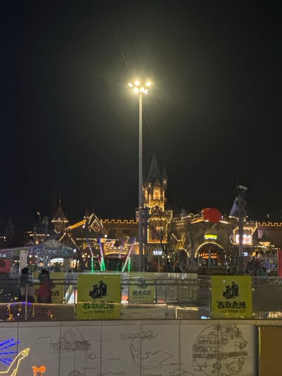 The image captures a vibrant night scene at what appears to be an amusement park or a themed entertainment area. The primary light source, a tall lamppost with multiple bright bulbs, is positioned on the left side of the frame, emitting a strong, starburst-like glow that illuminates the immediate surroundings and casts light across the dark sky.

Dominating the central and right portions of the image is a complex of buildings designed in a castle or fantasy village style. These structures are extensively decorated with warm, golden string lights that outline their architectural features, including turrets, battlements, and arched entrances. The lighting gives the buildings a festive and inviting appearance. Among these structures, what appears to be a brightly lit ride, possibly a swing ride or carousel, is visible with arms extending upwards, adorned with colorful lights in red, green, and blue. A large, round, red object, possibly a balloon or a significant light fixture, is suspended above one of the buildings.

In the foreground on the right, there is a barrier or wall. Several bright yellow rectangular banners are prominently displayed on this wall. Each banner features a cartoon character, which appears to be a stylized bear or panda, and includes black Chinese characters. The characters "熊出沒" (Xióng Chūmò), known as "Boonie Bears," are clearly visible on some of these banners, suggesting a tie-in with the popular Chinese animated series. Below these banners, the wall surface is patterned with a grid and adorned with various neon-like light patterns. These illuminated designs include outlines that resemble maps or playful illustrations. Near the bottom right corner, a glowing blue outline of a hand and the distinct "Adidas" logo rendered in light are also visible.

Several indistinct figures of people can be seen in the lower right part of the image, near the illuminated wall, indicating activity and attendance at the location. The overall atmosphere is lively, festive, and brightly lit against the dark backdrop of the night sky.