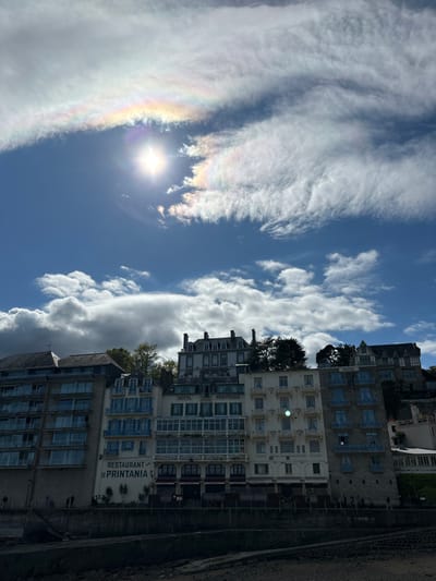 The image shows a row of buildings located in Dinard, France at the coordinates 48.636, -2.049. The buildings are all multi-story and made of stone, with a variety of architectural styles. The buildings are located on a waterfront, with a beach in the foreground. The sky is bright blue and filled with white clouds. There are a few people walking on the beach in the foreground.  The sun is shining brightly and there is a faint halo of light around it. The building in the center is labeled "Hotel Printania" above the entrance and "Restaurant Le Printania" below. The building to the right is labeled "Hotel" above the entrance. There is a faint rainbow-like effect in the clouds directly above the sun.  There are trees and foliage in front of the buildings.  There are shadows cast from the buildings and the trees onto the beach.