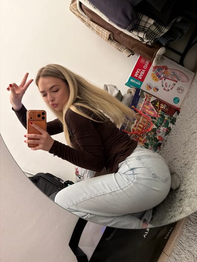 A woman poses in front of a round mirror, giving a peace sign with her left hand while holding a phone in her right. She has long blonde hair, light skin, and wears a dark brown long-sleeved shirt and light blue jeans. The background shows cluttered items like boxes (one labeled "Baby Gym Mat"), clothes, and a black bag. The setting appears to be a room with a carpeted floor.