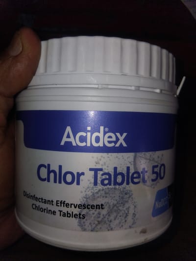 A white plastic container labeled "Acidex Chlor Tablet 50" is held in a person's left hand. The container has a blue label with white text. The text "Disinfectant Effervescent Chlorine Tablets" is visible below "Chlor Tablet 50". A blue rectangular sticker with "NaDCC" is on the lower right side of the container. The background is dark. The image was taken in Haveli Jhangranwali, Pakistan.