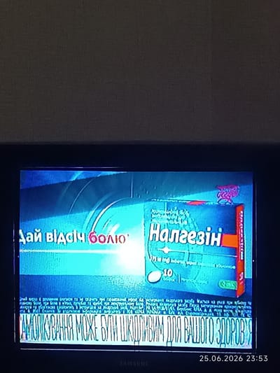 The image shows a television screen displaying what appears to be a program with on-screen text. The main subjects are text graphics and what seems to be a logo. The scene is set within a room, implied by the presence of the television.

The screen displays two prominent lines of text in Ukrainian. The top line reads "Дай відсіч болю" which translates to "Fight off the pain." Below this, the word "Налгезін" (Nalgezin) is prominently displayed, which is likely the name of a medication. To the right of "Налгезін," there is a pinkish-red graphic that could be a logo or an advertisement element. Below the main text, there is a smaller block of text, which is somewhat obscured and pixelated, but appears to be a disclaimer or additional information. The bottom line of large, stylized text reads "САМОЛІКУВАННЯ МОЖЕ БУТИ ШКІДЛИВИМ ДЛЯ ВАШОГО ЗДОРОВ'Я" which translates to "Self-medication can be harmful to your health."

In the lower right corner of the screen, a timestamp is visible: "25.06.2026 23:53," indicating the date and time. The overall impression is that of a medical advertisement or public service announcement related to pain relief and the dangers of self-medication, broadcast in the evening. The colors on the screen are predominantly blue and bright white for the text, with accents of red and pink in the graphics. The television itself appears to be an older model, possibly a CRT television, given the aspect ratio and some distortion of the image.