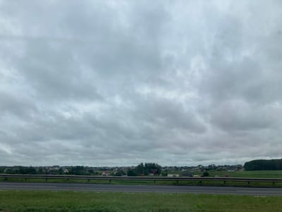 The image depicts a cloudy sky over a rural landscape in the village of Pekalin, within the Pekalinsky Selsoviet, Belarus. In the foreground, a highway or road runs horizontally across the frame, bordered by a grassy median strip on the left and a guardrail on the right. Beyond the guardrail, a green field stretches towards a village that is spread out on rolling hills. Numerous houses, varying in size and style, are visible among trees. The sky is overcast with heavy, grey clouds that dominate the upper half of the image. The overall impression is one of a subdued, possibly cool, day in a rural Belarusian setting.