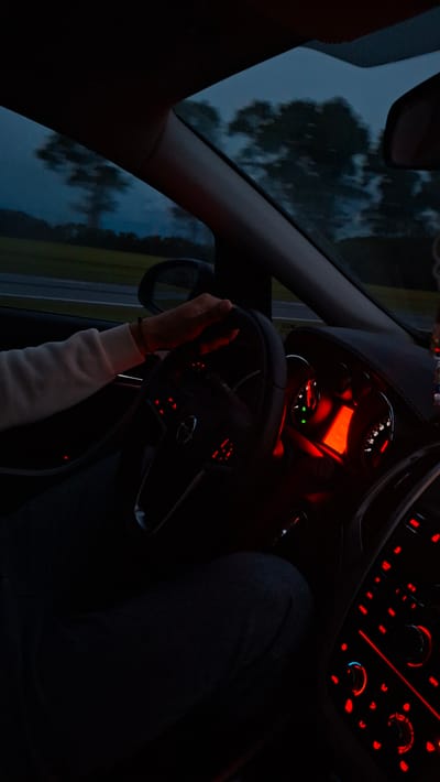 The image depicts the interior of a car at night in Самахвалавіцкі сельскі Савет, Belarus. The dashboard and steering wheel are visible, illuminated by red lights. A person wearing a white sweater is holding the steering wheel. The outside environment is visible through the car window.