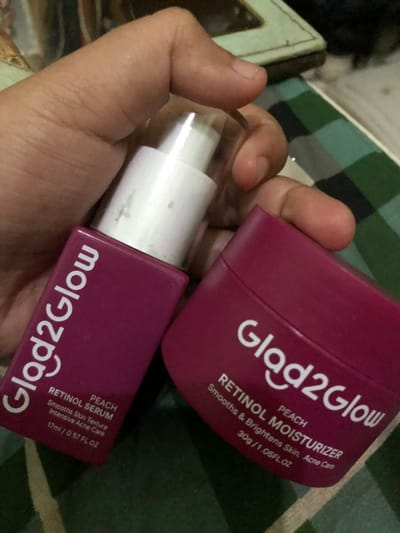 A human hand with light-to-medium skin tone is holding two magenta-colored cosmetic products in the city of Tlogo, Indonesia. The object on the left is a "Glad2Glow PEACH RETINOL SERUM" in a 17ml pump bottle with a clear cap, labeled for "Smooths Skin Texture Intensive Acne Care." The object on the right is a "Glad2Glow PEACH RETINOL MOISTURIZER" in a 30g jar, labeled for "Smooths & Brightens Skin, Acne Care." Both products feature white text on their labels. The background is out of focus, revealing a dark green and black plaid pattern, along with indistinct lighter and darker shapes.