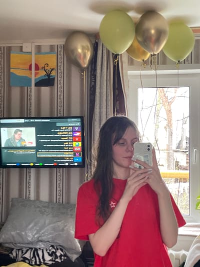 A young woman in a red shirt is taking a selfie with a smartphone in front of a window, likely indoors. To her left are several balloons in colors of gold and pale green. Behind her is a wall with wallpaper, a television displaying Russian text and images, and a silver pillow resting on a striped garment. Above the television is an abstract painting of a landscape with sun, sand, and a tree. The window behind the woman shows a tree and buildings outside. The location could potentially be in Ulukulevo, Russia, as indicated by the Russian text on the television.