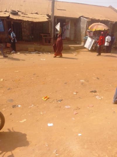 This image depicts a dusty street scene in Lapai, Nigeria, under bright daylight. The foreground is an unpaved, reddish-brown dirt road, heavily littered with various pieces of debris, including paper, plastic, and wrappers; one distinct yellow and red wrapper lies centrally.
In the mid-ground, a person in a full reddish-brown burqa or abaya walks from left to right, casting a shadow to their right. Behind them, along the right side, multiple individuals are gathered near a commercial establishment. A person in a red long-sleeved shirt stands near a vibrant orange and white striped market umbrella, which shades a display or stall. Two other individuals are partially visible behind them. A white rectangular sign, possibly a signboard, leans against an object near the umbrella.
On the left side of the scene, near a dilapidated building with a corrugated metal roof and a tire resting on it, a male figure in a light blue shirt and dark trousers stands with his back partially turned. Wooden benches or tables are visible outside this structure. A utility pole stands centrally, with a white triangular sign attached, pointing right. The buildings lining the street feature corrugated metal roofs and metal security gates, suggesting commercial premises. One building on the right has a partially visible white sign with red lettering ("AS"). A motorcycle wheel and its shadow are visible in the bottom-left corner of the frame.