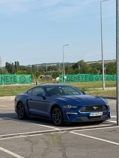 A dark blue Ford Mustang coupe is parked in an asphalt lot under bright daylight conditions in Kapitan Andreevo, Bulgaria. The vehicle is positioned facing forward and slightly to the left, with its front end near a vertical metal pole on the right side of the frame. The Bulgarian license plate "PB 9425 HA" is prominently displayed on the front bumper. The car features black multi-spoke alloy wheels. White painted lines delineate a parking space in the asphalt foreground, which also exhibits a wet or stained patch. In the midground, a long green mesh banner stretches horizontally, bearing the word "GOLDENEYE" repeated in black capital letters, interspersed with a circular target-like logo. Behind the banner, a grassy area contains various trees, shrubs, and large industrial-style wooden spools. A yellow rhombus-shaped road sign (priority road) is visible above the right side of the banner. Several tall, slender streetlights are present in the background, along with a distant power pylon on the far left. Rolling green hills form the horizon under a clear, light blue sky with minimal cloud cover.