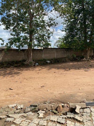A dirt road in Sabo, Nigeria, runs alongside a grey concrete block wall and under the shade of two trees. The ground is mostly bare dirt, transitioning on the right to broken grey concrete pavers. Trash is visible at the base of the wall. Above, the sky is partly visible through the trees, marked by pale blue and white clouds.