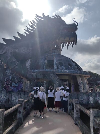 A group of approximately ten individuals, primarily women dressed in white tops and dark skirts or pants, stands on a concrete walkway with low railings, observing a large, derelict dragon-shaped concrete structure. This structure is located in Kim Long Ward, Vietnam, and consists of a colossal, weathered dragon sculpture integrated with a dome-like building. The dragon's head, with an open mouth and prominent fangs, faces right, silhouetted against a bright, cloudy sky with strong sunlight causing lens flare. Its body wraps around the dome, which features circular and rectangular openings. The entire edifice shows signs of significant decay, including exposed concrete and extensive multi-colored graffiti. Several individuals in the group hold smartphones, appearing to photograph the structure. Shadows are cast on the walkway where the people stand. Overgrown vegetation is visible around the base of the structure and in the background.