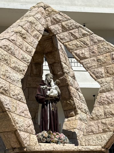 A statue of Saint Anthony of Padua holding the infant Jesus stands within an angular stone archway. The statue depicts a man in a brown Franciscan habit, with a rosary around his neck. He cradles a child in his left arm and holds a book in his right. A floral arrangement sits at the base of the statue. The archway is constructed of rough-hewn, light-colored stone blocks. Behind the archway, a white building with a barred window can be seen. The scene is lit by natural daylight, casting shadows on the stone. This is located in Jezzine, Lebanon.