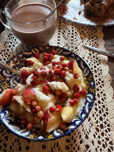The image shows a rustic, dimly lit scene, likely set in Voeikovo, Russia, on a wooden table covered with a cream-colored lace doily. In the foreground, a blue and yellow patterned ceramic plate holds a serving of what appears to be a porridge or rice pudding, topped with sliced apples and numerous small, red wild strawberries. A fork rests in the dish. To the left, a clear glass pitcher contains a dark, frothy beverage, likely hot chocolate. In the background, another plate holds a slice of pie or cake, partially visible. The overall impression is one of a comforting, home-style meal. There are no people visible in the image.