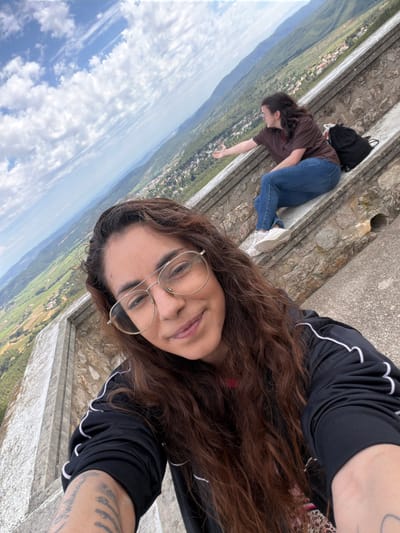 A selfie taken by a woman with brown curly hair and round glasses. She is wearing a black athletic jacket with white trim. The photograph is taken on a high, stone ledge. In the background, another woman is lying down on the stone, reaching out with her right arm. She is wearing jeans and a brown shirt. A black backpack sits next to her. Below the ledge, the landscape of Torrelles de Foix, Spain, is visible with fields, buildings, and mountains. The sky is blue with scattered clouds.