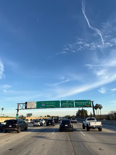 The image shows a congested highway scene under a clear blue sky with wispy clouds. Traffic is bumper-to-bumper, with a mix of cars and trucks visible. Overhead green highway signs indicate directions: South to LAX Airport and Long Beach via the 405, Slauson Avenue/Marina del Rey via the 90, and Washington/Culver Boulevards at the next exit. Additional signs point out the carpool lane.