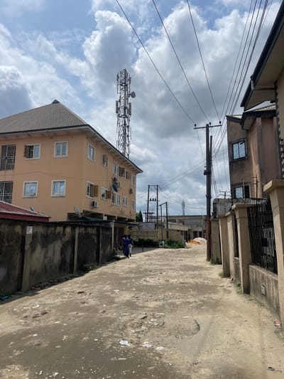 The image captures a daytime scene in Alozo, Nigeria, portraying a street or alleyway flanked by urban buildings under a partly cloudy sky. The foreground is an unpaved, dusty dirt path, showing wear and scattered debris. Concrete walls line the path, separating it from the adjacent properties.

On the right, a prominent multi-story building with a light brown/yellow facade features numerous windows, some equipped with security grills and several external air conditioning units. Two small satellite dishes are also mounted on its exterior. Behind this building, a tall, metal lattice communication tower, adorned with various antennas and equipment, stands against the backdrop of the sky. To the left, a darker, older-looking building is partially visible with some windows.

The sky is a mix of bright blue patches and significant white and gray cloud cover. A dense network of electrical and communication wires stretches across the frame, supported by utility poles, one of which is prominent in the middle distance.

A person dressed in blue attire is partially visible in the mid-ground, standing near the yellow building, close to a dividing wall. Other figures can be vaguely seen further down the path. The scene depicts a typical day in a residential or mixed-use area, characterized by visible infrastructure and local architecture. No legible text is visible in the image.