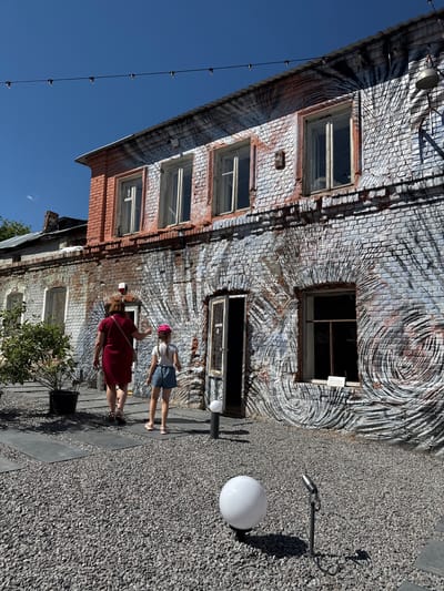 In Yelets, Russia, an adult woman in a red dress and a young girl wearing a pink hat and blue shorts walk on a gravel path towards a brick building. The building features a distinctive mural on its exterior walls, with a pattern resembling concentric circles and brush strokes. The mural is painted over aged bricks, some of which are red and others are a lighter, weathered hue. The building has multiple windows, some of which are open. To the left of the building, a potted plant sits near the path. The ground is covered in dark gray gravel, interspersed with dark gray rectangular stepping stones. In the foreground, a white globe-shaped lamp stands on a short black pole, and a small, dark spotlight on a stand is also visible. Overhead, a string of bare light bulbs hangs against a clear blue sky. The scene appears to be an outdoor courtyard area, possibly a public space or a commercial establishment, given the art on the building and the decorative lighting.