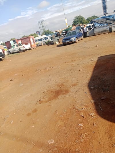 An open, dusty lot contains several vehicles including a white pickup truck and a blue sedan, parked amidst scattered debris. In the background, a white van is visible along with a billboard structure. Some people are gathered near a covered structure. The foreground is dominated by dry, reddish-brown soil with patches of darker stains. Shadows are visible on the right side of the image, adding depth to the scene. The sky is partly cloudy.