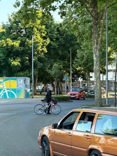 In Cleves, Germany, on a paved street bordered by trees and buildings, a man in a black shirt and pants walks a bicycle with a red frame and black accessories. He is positioned mid-street, near a yellow and green bike rack facility. To his right, a shiny orange station wagon is partially visible in the foreground, and further down the street, a small red car is driving away from the camera. A tall lamppost stands to the right of the man, and other trees and streetlights are scattered throughout the scene. The sky is clear and blue.