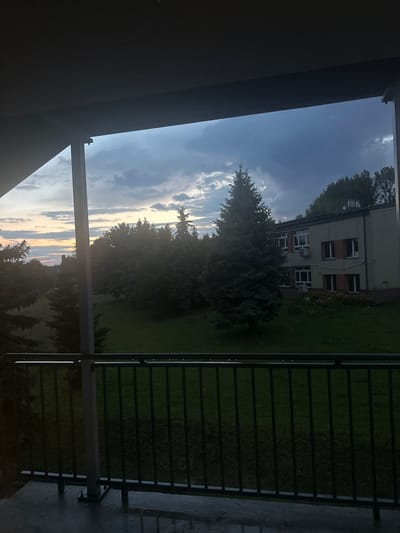 The image depicts an outdoor scene observed from a covered balcony or patio in Giebułtów, Poland, likely during twilight or early evening. The immediate foreground shows a concrete floor, appearing wet from recent rain, reflecting light subtly. A dark metal railing with vertical balusters and a horizontal top rail extends across the lower portion of the frame. To the left, a vertical support pillar for the overhead structure is visible.
Beyond the railing, a large, well-maintained grassy area forms the midground. Several mature trees, primarily conifers with dense foliage, are scattered across this green space, with a particularly prominent evergreen tree situated centrally. A cluster of deciduous trees is visible behind these, creating a partial treeline.
In the background, to the right, a two-story building with a light-colored facade is visible. It features multiple rectangular windows, some of which have closed, reddish-brown external shutters. Two white external air conditioning units are mounted below windows on the ground floor. A small garden bed with some visible flora is located near the building's base. Further in the background, a denser line of trees suggests a slightly elevated terrain or wooded area.
The sky above is characterized by dark, heavy clouds, with lighter, more illuminated sections along the horizon on the left, indicative of a sunset or sunrise. No individuals or discernible actions are present within the frame. The overall environment suggests a quiet, possibly residential or institutional setting.