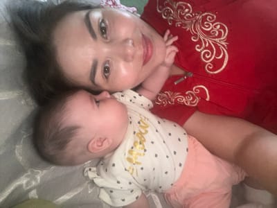A close-up, overhead shot shows a woman and a baby lying down, likely on a bed or sofa. The woman is on the right, wearing a red zippered jacket with ornate gold embroidery. Her dark hair is pulled back. She is smiling and looking at the camera. Her left hand rests on the baby's head. The baby is on the left, facing away from the camera, with their head turned slightly towards the woman. The baby is wearing a white onesie with a pattern of dark gray dots and the word "meow" in gold lettering. The baby is also wearing light pink shorts. The scene appears to be indoors, with a soft, diffused light suggesting it could be daytime. The overall mood is warm and affectionate, capturing a moment of closeness between mother and child. The visible text "meow" is on the baby's onesie.