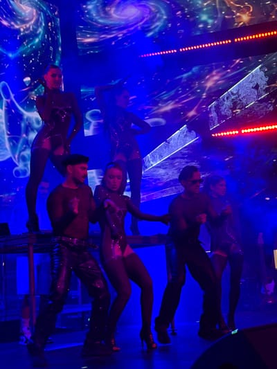 This image captures a performance on a stage bathed in vibrant blue and purple lighting, with a cosmic-themed backdrop featuring nebulae and starry effects. Four women in form-fitting, dark, jeweled costumes are dancing, their attire featuring cutouts and sheer elements. Three men are also on stage. One man, wearing a black beret and a textured black shirt with shiny black leather pants, stands in the foreground, seemingly interacting with the performers. Another man, wearing sunglasses and a dark, sheer long-sleeved shirt with black pants, is positioned to the right of the beret-wearing man. A third man is partially visible in the background, seated behind what appears to be DJ equipment. Red LED lights run along the upper edges of the stage structure. The scene suggests a live musical or performance event, possibly in a club or concert venue. The image does not provide any specific information regarding the location being Tsilkani, Georgia.