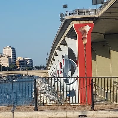 The image depicts a concrete vehicular bridge, identified as Ponte Risorgimento, spanning the Fiume Pescara in Pescara, Italy. The scene is viewed during daylight from the riverbank, looking towards the bridge's underside and piers.

The bridge's support structures are the primary subjects. The closest pier on the right is painted a solid red, featuring a prominent, vertically oriented, stylized white form resembling a calla lily or an elongated drop, with subtle brown internal striations. To the left, a series of adjacent concrete piers display large-scale, abstract geometric murals in black, white, and grey, with occasional red accents, forming repeating patterns along the bridge's length. The underside of the bridge deck is visible above the piers, constructed of light grey concrete.

In the midground, the Fiume Pescara appears as a calm, blue body of water reflecting the sky. Several small boats are partially visible moored in the river further downstream. Beyond the river, the urban landscape of Pescara is discernible, comprising several multi-story buildings of varying heights and pale colors, consistent with residential or commercial structures. A lower, smaller bridge is also visible in the distance across the river.

In the foreground, a dark metal railing with vertical bars and a top rail extends horizontally across the lower portion of the frame, acting as a barrier between the viewer's position and the river. Faint graffiti is present on the lower section of the railing. Below the railing, a strip of dry, light-brown earth or gravel transitions to a paved surface, which includes a curb and a visible storm drain grate. The sky is clear and light blue, indicating fair weather conditions. No individuals are present in the depicted scene.