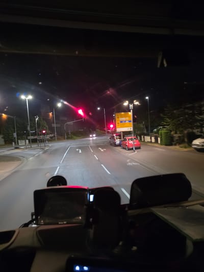 The image is a night-time, interior, forward-facing view from a vehicle's dashboard, likely a truck, looking out through the windshield. The scene depicts a street intersection in Flörsheim, Germany. Several streetlights illuminate the road and surrounding area. A traffic light in the distance is illuminated red, indicating a stop. Several cars are visible on the road: a red hatchback, a silver van, and other vehicles further down the street. A yellow directional sign with German text is mounted on a pole. The dashboard features two electronic devices, one with a screen, and a clipboard with papers. The overall environment appears to be a suburban or semi-urban area of Flörsheim.
