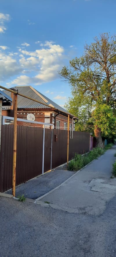 The image captures a residential street scene in Shakhty City District, Russia. A red brick house with a grey corrugated metal roof and white accents is visible behind a tall, dark brown metal fence. A satellite dish is mounted on the side of the house. The fence is supported by yellow metal posts. A narrow, paved walkway runs alongside the fence, with green vegetation growing along its edge. A large, mature tree with a thick trunk and green foliage stands to the right of the house, casting shadows. The sky above is blue with scattered white clouds, illuminated by what appears to be late afternoon or early evening sunlight. The road in the foreground is asphalt with visible cracks and patches.