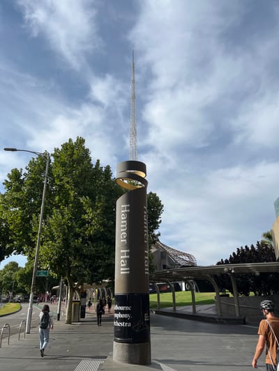 This daytime photograph captures a street-level view of a cultural precinct in Southbank, Australia, centered around the distinctive signage for Hamer Hall. The image is composed with a blend of architectural elements, natural foliage, and people.
A prominent feature is a tall, slender spire-like structure, likely the Arts Centre Spire, which ascends high into a partly cloudy sky. Near its base, the spire interacts with a large cylindrical sign. This sign, dark brown or grey, features a unique golden, ribbon-like structure wrapped around its upper portion. The words "Hamer Hall" are clearly and boldly displayed in light-colored text twice on the sign, along with "Melbourne Symphony Orchestra" in smaller letters below.
To the right of the sign, the leafy green canopy of large trees fills a significant portion of the midground. The ground is paved, indicating a public walkway or plaza, and several metal bike racks are visible, with multiple bicycles parked. People are present in the scene; a person wearing a brown shirt and a cycling helmet stands with their bicycle on the right. Another individual, carrying a backpack, is seen near the bike racks to the left of the sign.
In the background, hints of modern architecture are visible, including a building with an interesting, possibly patterned or lattice-like roof structure. The sky is bright with scattered white clouds, suggesting clear and pleasant weather. The scene portrays a typical day in a busy urban cultural hub.